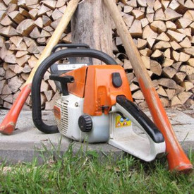 Vor einem Stapel von gespaltetem Brennholz steht eine Motorsäge auf dem Boden. Dahinter lehnen eine Spaltaxt und ein Spalthammer schräg an einem Holzklotz.